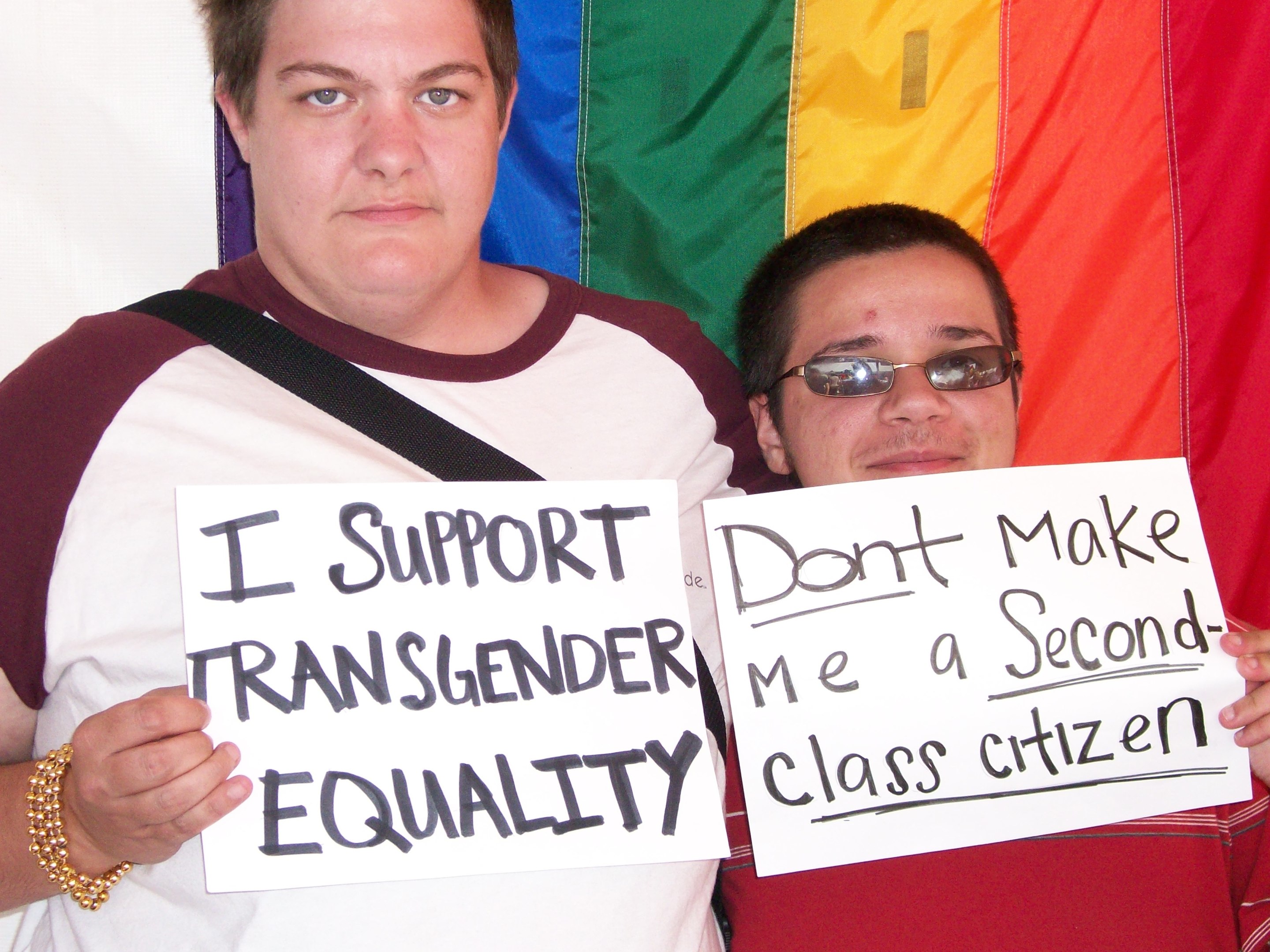 Two individuals pose in front of rainbow banner with signs reading "I Support Transgender Equality" and "Don't Make Me a Second-Class Citizen."
